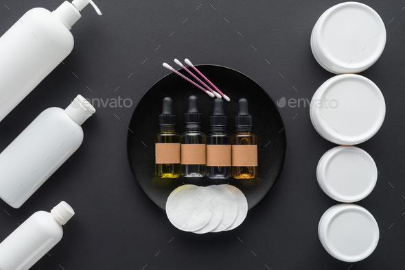 top view of bottles of cream, natural oils and cosmetic pads isolated on black, beauty concept