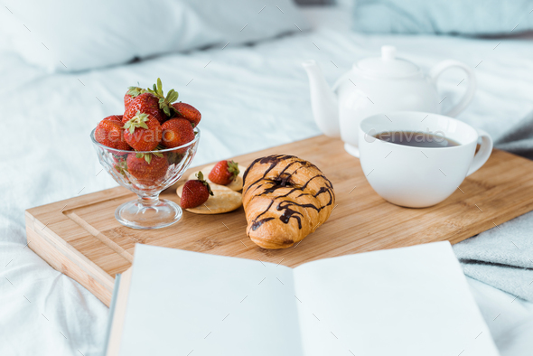 appetizing breakfast of strawberries, croissant and coffee on wooden tray on bed