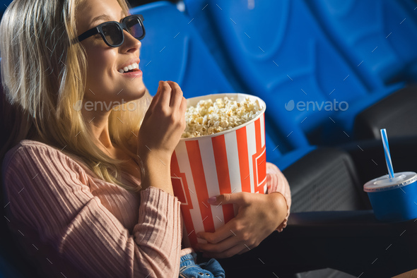 side view of smiling woman in 3d glasses with popcorn watching film alone in cinema