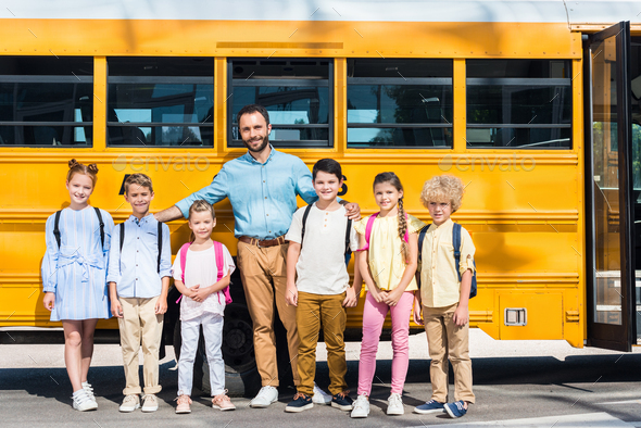 schoolchildren and teacher standing together in front of school bus and looking at camera