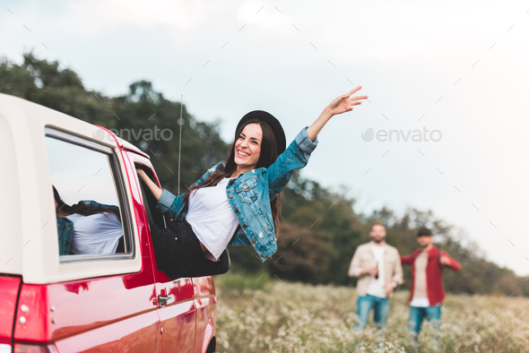 young woman outstretching from car window and raising hand while men standing blurred on background