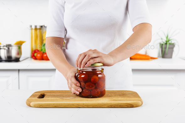 cropped image of woman opening glass jar with preserved tomatoes in light kitchen