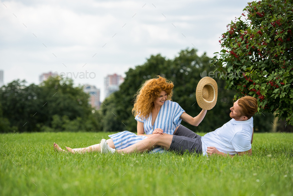 smiling redhead woman trying to putting on own straw hat on boyfriend head on grass in park