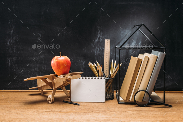 close up view of apple on wooden toy plane, notebook, books and pencils on tabletop with empty