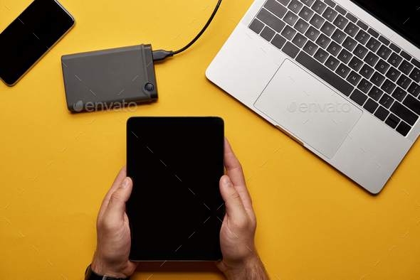 cropped shot of man using tablet on yellow surface with laptop and portable hdd
