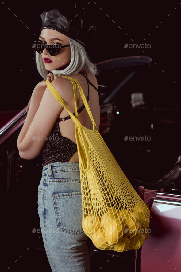 stylish blonde girl in sunglasses and beret holding string bag with lemons standing near vintage car