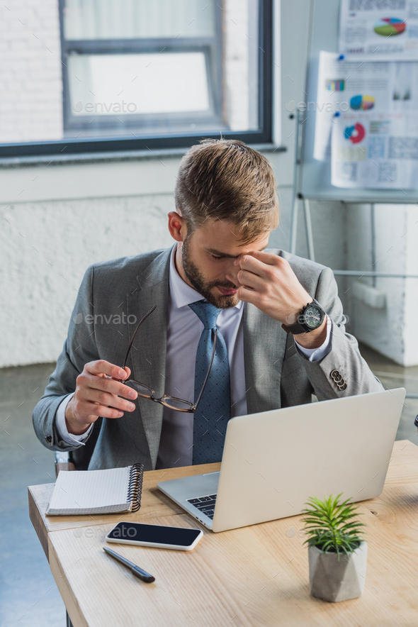 high angle view of tired young businessman holding eyeglasses and rubbing nose bridge while using
