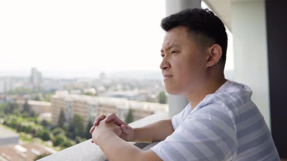 A Young Asian Man Looks Thoughtfully Around on an Apartment Balcony in an Urban Area  Closeup alt