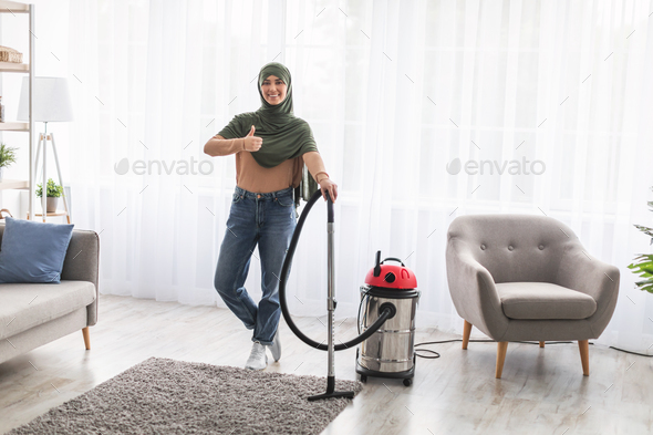 Muslim woman cleaning carpet with vacuum cleaner, showing thumbs-up