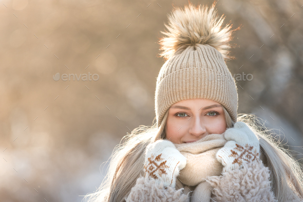 Winter outdoor portrait of young woman in warm hat, knitted mittens and scarf walking in snowy park