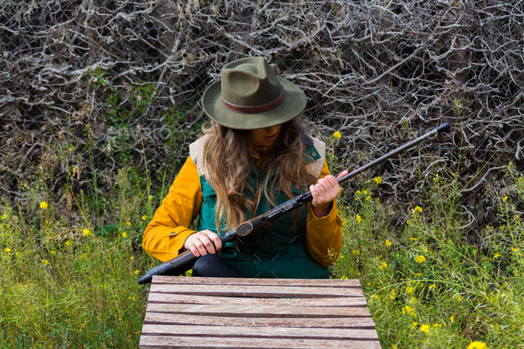 Blonde scout girl in green vest and green hat preparing weapon for hunting.