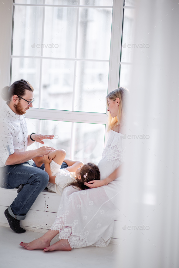 Little girl laughs lies on the lap of pregnant mother the father tickles daughter. Happy family