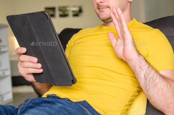 Casual young man looking at digital tablet and saying hello in the living room