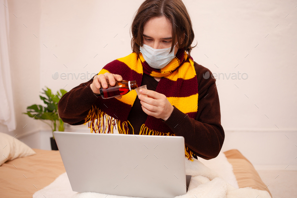 A sick man sits at a laptop wearing a mask. Coronavirus, covid, home quarantine. Man pours himself