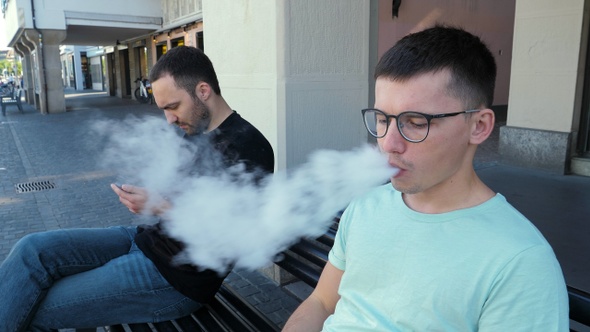 Handsome hipster in glasses smoking vape on a bench next alt