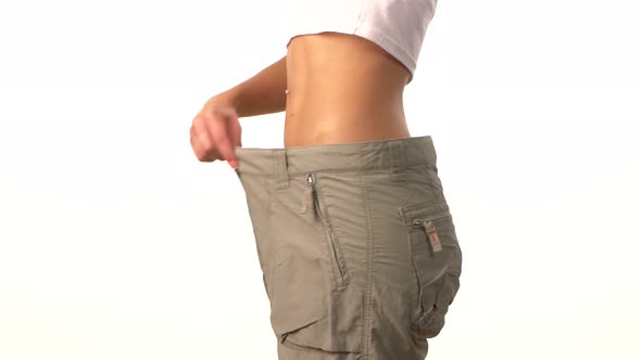 Slim Girl in Big Trousers, Showing Her Lose Weight, Hand on Her Waist, on White alt