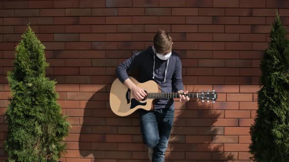 Young Man with Medicinal Mask on Face Is Playing at the Guitar Outside. alt