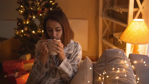 Camera Zooming to Happy Dreaming Girl in Pajama Sitting on Couch with Lights Drinking Hot Chocolate alt