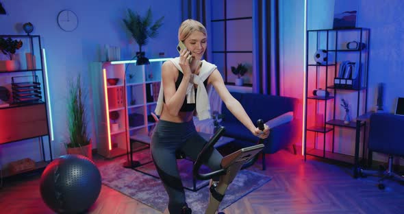 Young Woman in Training Clothes Turning the Pedals on Stationary Bike and Talking on Phone alt
