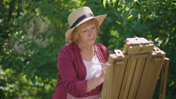 Thoughtful Senior Woman Painting in Sunshine Outdoors and Smiling Looking at Camera alt