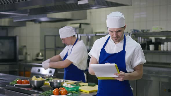 Professional Caucasian Male Chef Checking Orders Writing with Pen and Looking at Camera Smiling alt