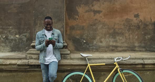 Excited Afro American Man Using His Smartphone To Play Online Games While Standing Leaned To Old alt