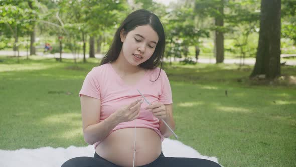 Happiness young asian woman pregnant sitting doing knit hat with for relaxation in the park. alt