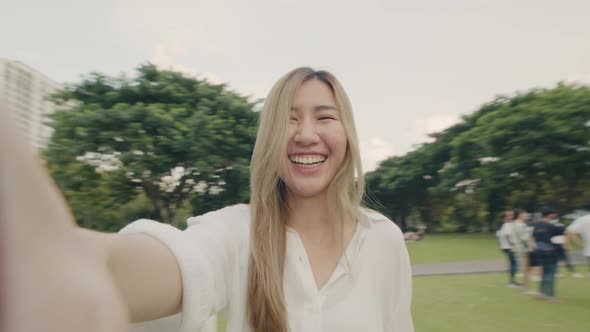 Happy beautiful young Asian woman taking selfies on a smartphone at a public park.