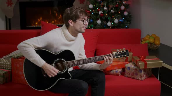 Guy is playing Christmas song on guitar in a decorated Christmas livingroom.