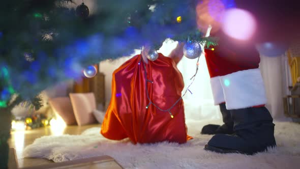 Legs of Unrecognizable Santa Walking to Decorated Christmas Tree in Living Room Putting Gift Box on alt