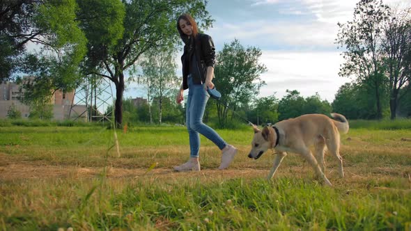 Wide Steadicam Shot of Attractive Girl in Jeans in Leather Jacket with Her Dog in the Park. alt