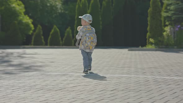 Lost Baby Boy Child in Cap Walking Street with Backpack and Teddy Bear in Hands alt