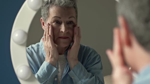 An Elderly Graying Woman Carefully Examines Her Reflection in the Mirror Lightly Touching the