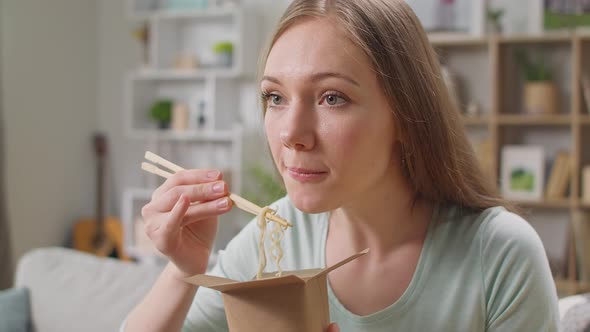 Young Woman Eating Noodles From a Box with Chopsticks at Home on a Couch alt