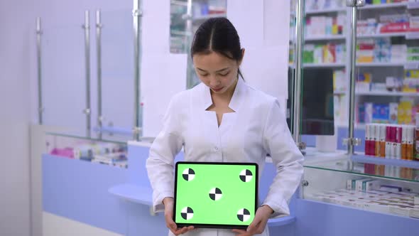 Young Asian Woman Posing with Green Screen Digital Tablet at Cash Desk in Pharmacy alt