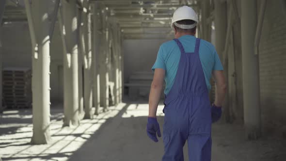 Back View of Adult Caucasian Man in Uniform and Helmet Walking on Construction Site in Sunlight alt
