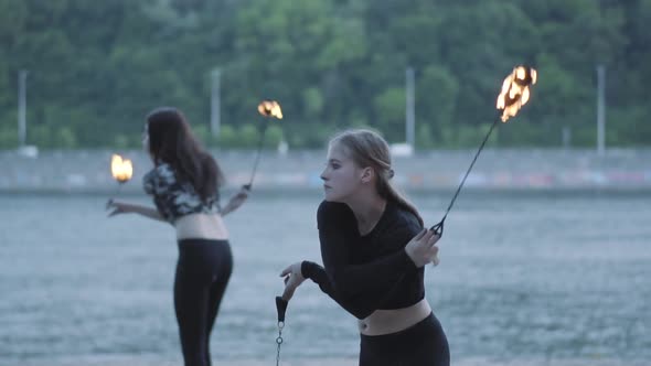 Two Young Women Performing a Show with Flame Balls Standing on the Riverbank alt