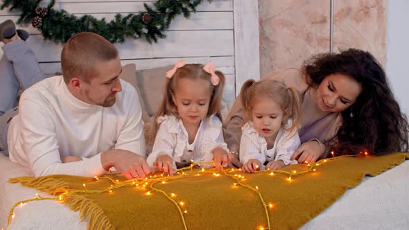 Friendly Family Lying on Soft Bed Decorated Christmas Garland Lights in Cozy Bedroom. Mother and