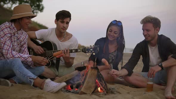 Group of Young and Cheerful People Sitting By the Fire on the Beach in the Evening Grilling Sausages alt