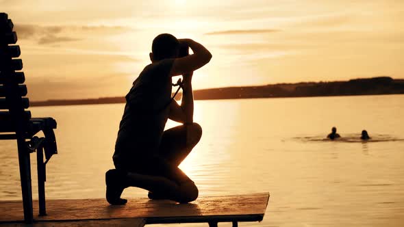 A man in shorts and a t-shirt is squatting and holding a camera focused on the sunset alt