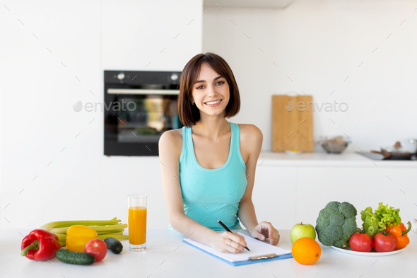 Happy fitness lady in sportswear writing daily ration diet or healthy recipe, standing in kitchen