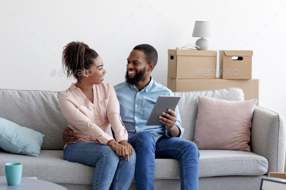 Happy young black male and female relax on couch using tablet for online buying furniture
