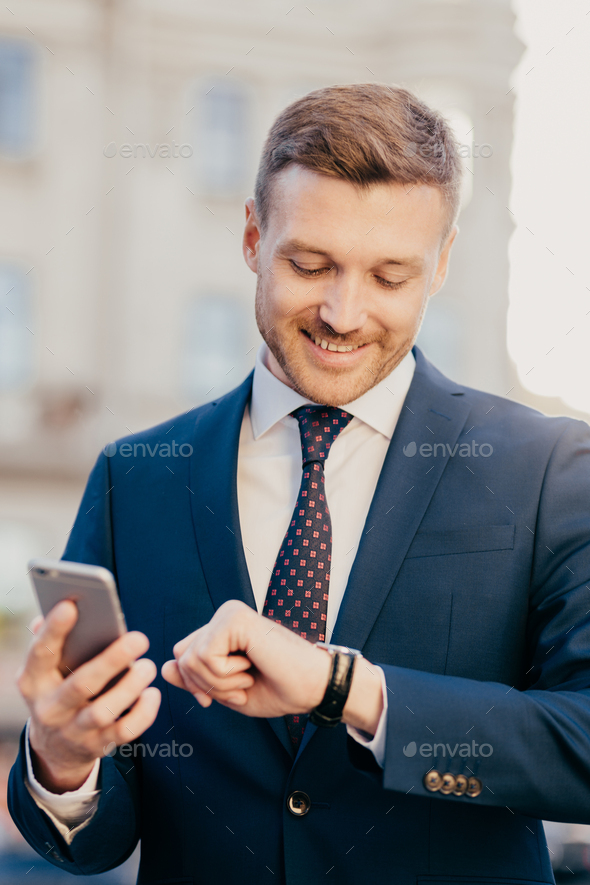 Male lawyer waits for client outdoor, looks at wristwatch, holds smartphone as waits for call