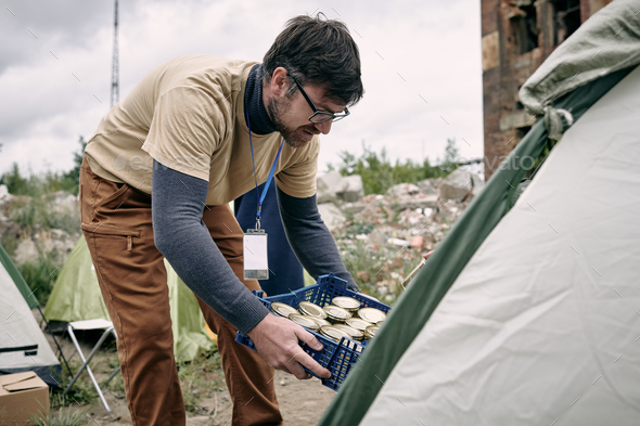 Male volunteer with box of canned food bending by tent