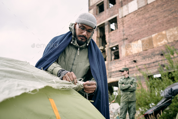 Male refugee pitching tent against half ruined building