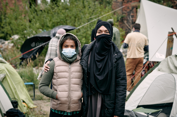 Young woman in hijab and her daughter standing against tent camp