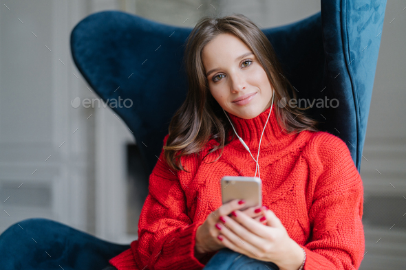 Satisfied female melamine enjoys favourite music, listens audio record, dressed in red sweater