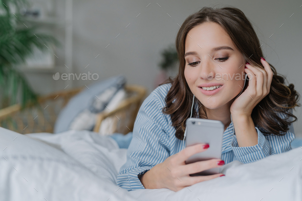 Beautiful woman listens music, makes video call, lies in comfortable bed in morning
