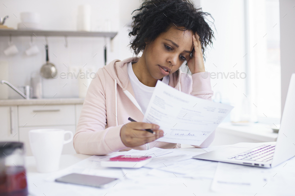 Young african woman feeling stressed while managing family budget at kitchen table