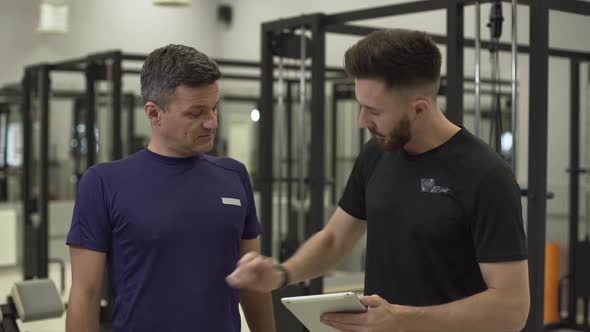 Professional Fitness Trainer with a Tablet in Hands Explaining the Plane of Training for a Sportsman alt